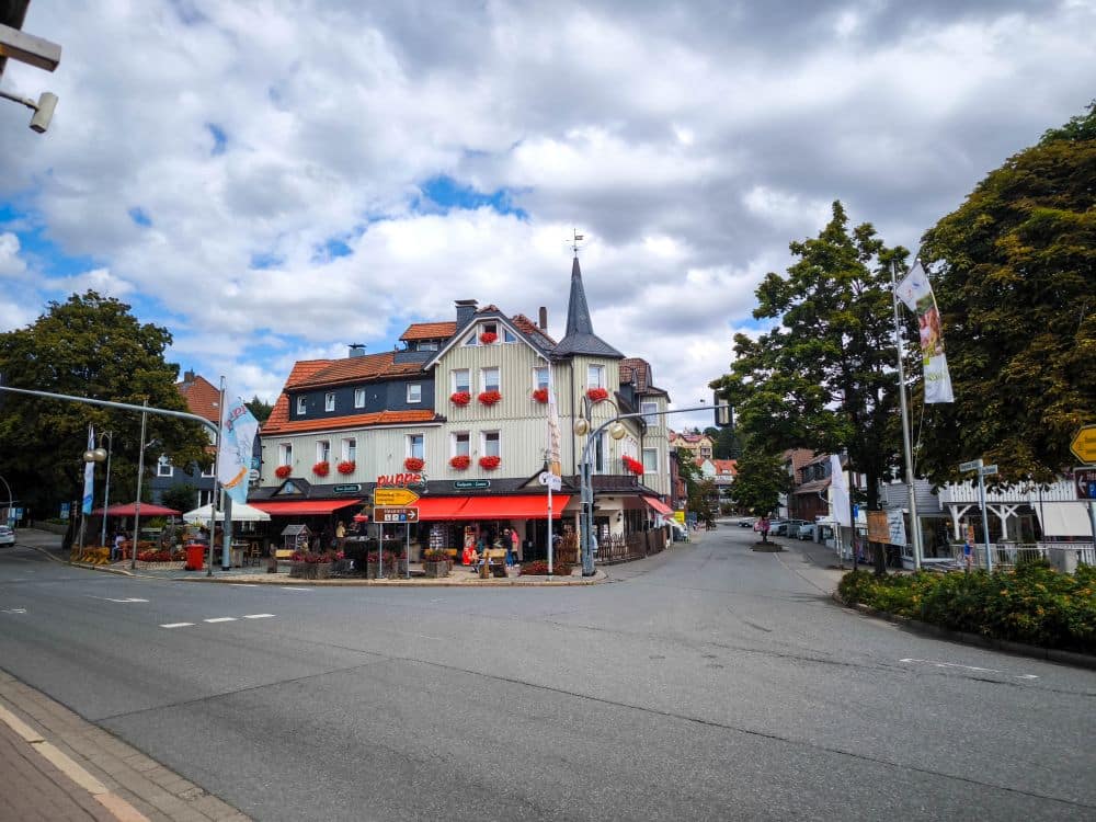 Ein charmantes Eckhaus mit einem spitzen Turm und roten Blumenkästen steht an einer Kreuzung in der ruhigen Stadt Braunlage. Der Himmel ist teilweise bewölkt, und Bäume und Straßenschilder säumen die ruhigen Straßen.