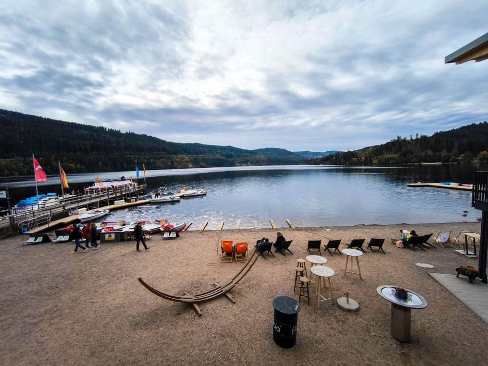 Eine ruhige Szene am Titisee mit Booten am Steg, Menschen, die sich auf einem Sandstrand mit Stühlen und Tischen entspannen, umgeben von bewaldeten Hügeln unter einem bewölkten Himmel.