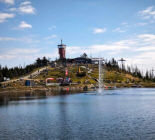 Menschen versammeln sich an einem See in Braunlage, umgeben von den malerischen Harzer Hügeln und Bäumen. Der rot-weiße Wurmbergturm steht oben auf dem Hügel, Gebäude und ein Skilift sind unter einem teilweise bewölkten Himmel zu sehen.