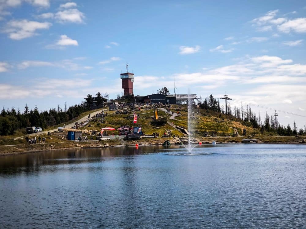 Menschen versammeln sich an einem See in Braunlage, umgeben von den malerischen Harzer Hügeln und Bäumen. Der rot-weiße Wurmbergturm steht oben auf dem Hügel, Gebäude und ein Skilift sind unter einem teilweise bewölkten Himmel zu sehen.