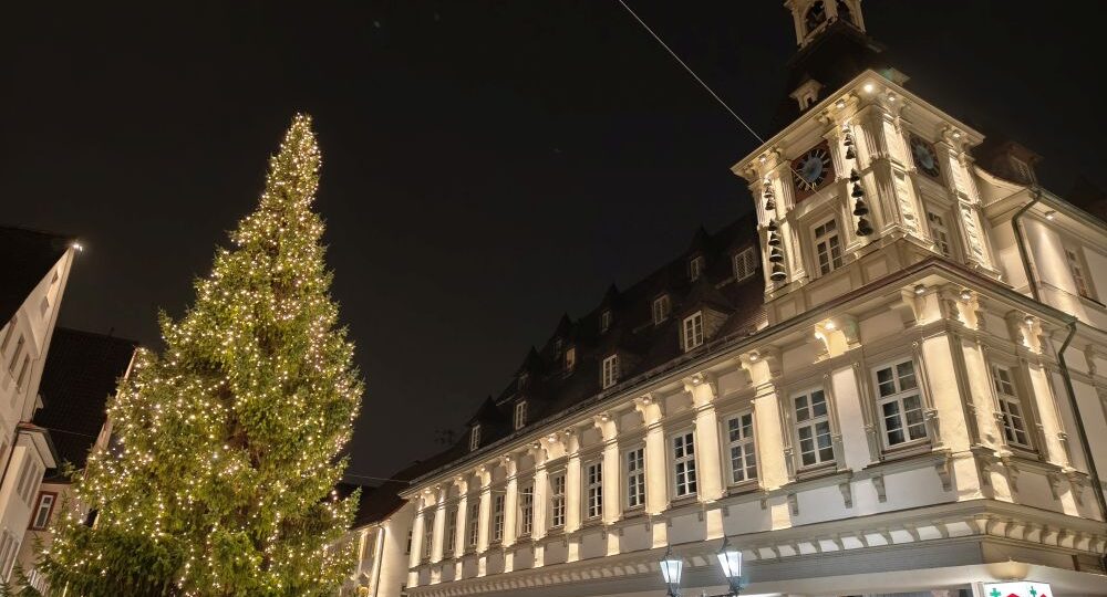 Ein großer, geschmückter Weihnachtsbaum mit Lichtern steht vor einem beleuchteten, historischen Gebäude mit einem Uhrenturm bei Nacht. Im Erdgeschoss sind Geschäfte mit beleuchteten Schildern zu sehen.