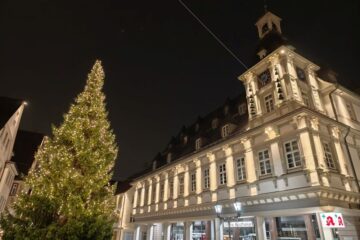 Ein großer, geschmückter Weihnachtsbaum mit Lichtern steht vor einem beleuchteten, historischen Gebäude mit einem Uhrenturm bei Nacht. Im Erdgeschoss sind Geschäfte mit beleuchteten Schildern zu sehen.