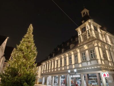 Ein großer, geschmückter Weihnachtsbaum mit Lichtern steht vor einem beleuchteten, historischen Gebäude mit einem Uhrenturm bei Nacht. Im Erdgeschoss sind Geschäfte mit beleuchteten Schildern zu sehen.