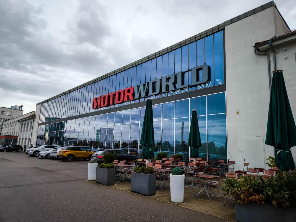 Außenansicht des MOTORWORLD-Gebäudes in Böblingen mit großen Glasfenstern, in denen sich der Himmel spiegelt, Sitzgelegenheiten im Freien mit Tischen und Stühlen, Topfpflanzen, geschlossenen Sonnenschirmen und parkenden Autos an der Seite.
