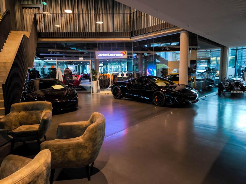 In einem Ausstellungsraum in Böblingen werden schwarze Luxussportwagen, darunter auch McLaren-Modelle, in hellem Licht präsentiert. Im Vordergrund stehen moderne Stühle, während eine Glaswand mit einem McLaren-Schild die elegante Atmosphäre vervollständigt.