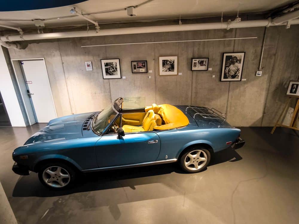 Ein klassisches blaues Cabriolet mit gelbem Interieur steht in einer modernen Böblinger Galerie mit grauen Betonwänden, gerahmten Schwarz-Weiß-Fotos und einer geschlossenen weißen Tür im Hintergrund.