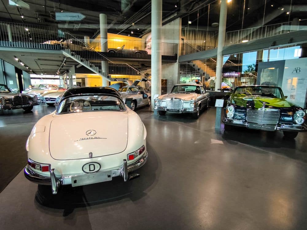 In einem modernen, lichtdurchfluteten Ausstellungsraum in Böblingen mit Glaswänden und einem Zwischengeschoss, in dem weitere Fahrzeuge zu sehen sind, wird eine Sammlung klassischer Mercedes-Benz Fahrzeuge präsentiert.