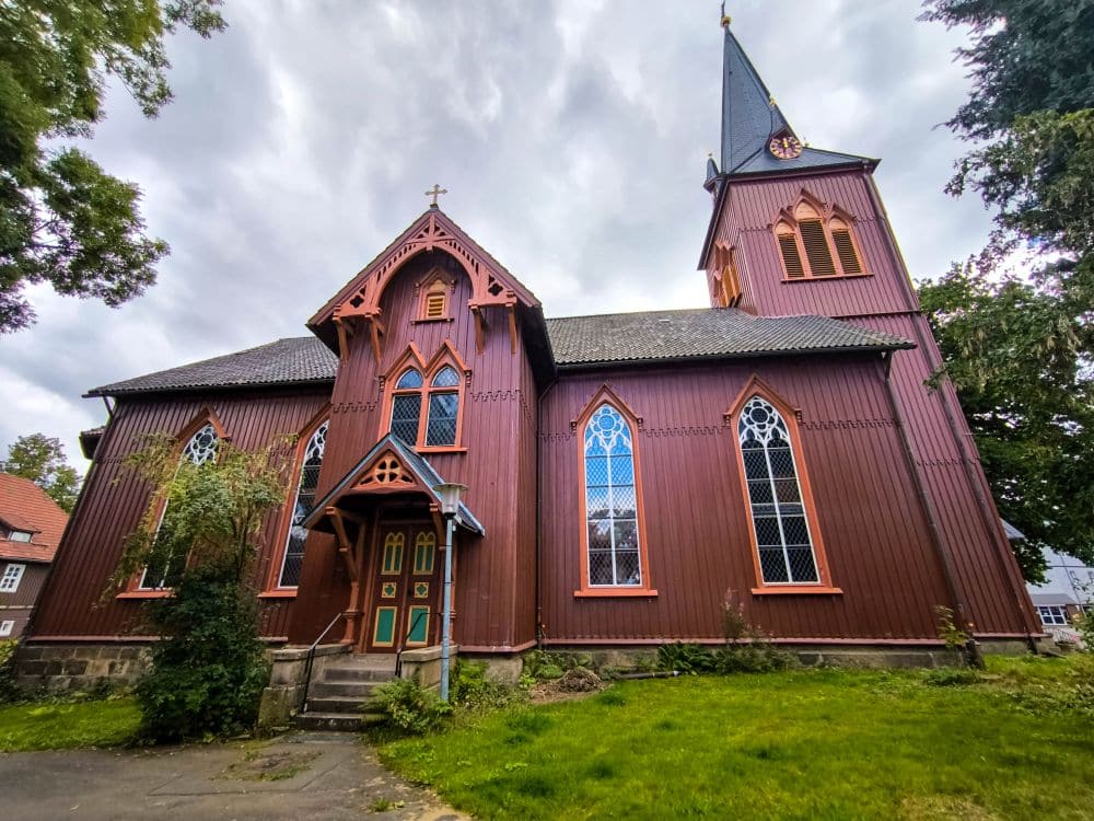 Eine rote Holzkirche mit Spitzbogenfenstern, einem steilen Dach und einem hohen Kirchturm steht inmitten von grünem Gras und Bäumen unter einem wolkenverhangenen Himmel in Braunlage. Stufen führen zu einer Eingangstür mit Zierleisten.