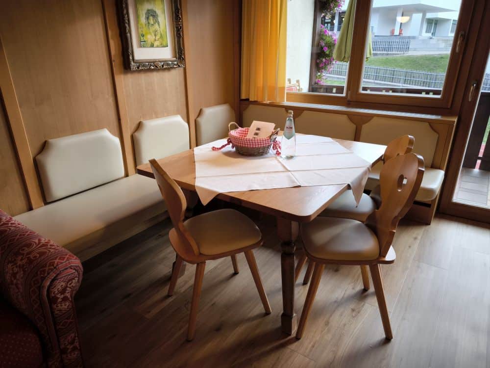 Eine gemütliche Essecke von Kastel besteht aus einem Holztisch, zwei Holzstühlen und einer Bank mit weißen Kissen. Ein Korb, eine Flasche und eine Speisekarte stehen auf dem Tisch, während große Fenster mit gelben Vorhängen den Raum in natürliches Licht tauchen.