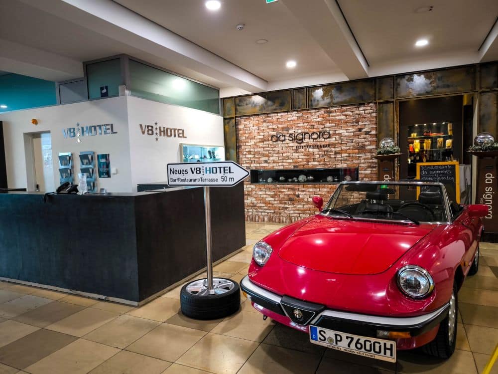 Ein leuchtend rotes Oldtimer-Cabrio ist in der Nähe der Rezeption einer modernen Hotellobby in Böblingen geparkt, mit einer Backsteinwand und der Aufschrift V8 HOTEL. Ein Hinweisschild für das Hotel steht neben dem Auto.