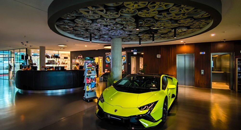 Ein leuchtend gelber Lamborghini-Sportwagen parkt in einer modernen, stilvollen Lobby in Böblingen mit einer runden Deckengestaltung, die Filmrollen zeigt. Im Hintergrund befinden sich ein Empfangstresen, ein Zeitschriftenständer und ein Aufzug.