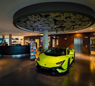 Ein leuchtend gelber Lamborghini-Sportwagen parkt in einer modernen, stilvollen Lobby in Böblingen mit einer runden Deckengestaltung, die Filmrollen zeigt. Im Hintergrund befinden sich ein Empfangstresen, ein Zeitschriftenständer und ein Aufzug.