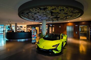 Ein leuchtend gelber Lamborghini-Sportwagen parkt in einer modernen, stilvollen Lobby in Böblingen mit einer runden Deckengestaltung, die Filmrollen zeigt. Im Hintergrund befinden sich ein Empfangstresen, ein Zeitschriftenständer und ein Aufzug.