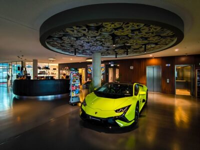 Ein leuchtend gelber Lamborghini-Sportwagen parkt in einer modernen, stilvollen Lobby in Böblingen mit einer runden Deckengestaltung, die Filmrollen zeigt. Im Hintergrund befinden sich ein Empfangstresen, ein Zeitschriftenständer und ein Aufzug.