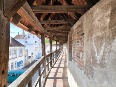Überdachter Gehweg mit Holzbalken und Ziegeldach, der entlang der historischen Stadtmauer verläuft, mit Blick auf eine Straße mit Gebäuden und einem geparkten türkisfarbenen Lieferwagen.