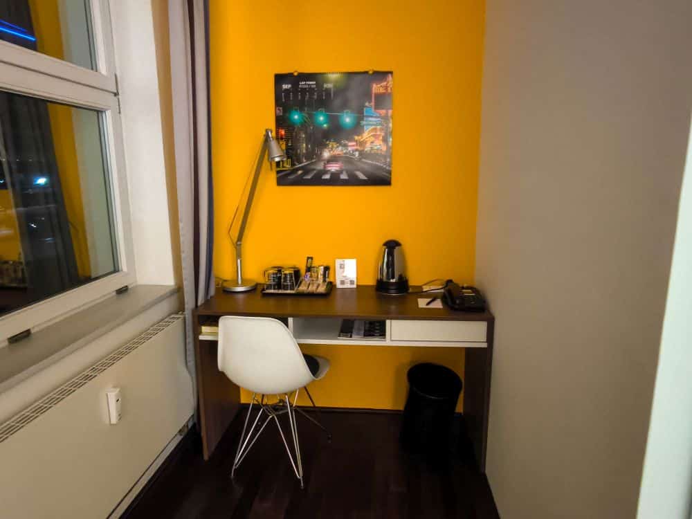 Ein kleiner Schreibtisch mit einem weißen Stuhl, einer Schreibtischlampe, einem Schachbrett, einem Wasserkocher und einem Telefon steht vor einer gelben Akzentwand in einem gemütlichen Böblinger Zimmer, über dem ein Stadtbild zu sehen ist. Links befindet sich ein Fenster und unter dem Schreibtisch ein Mülleimer.