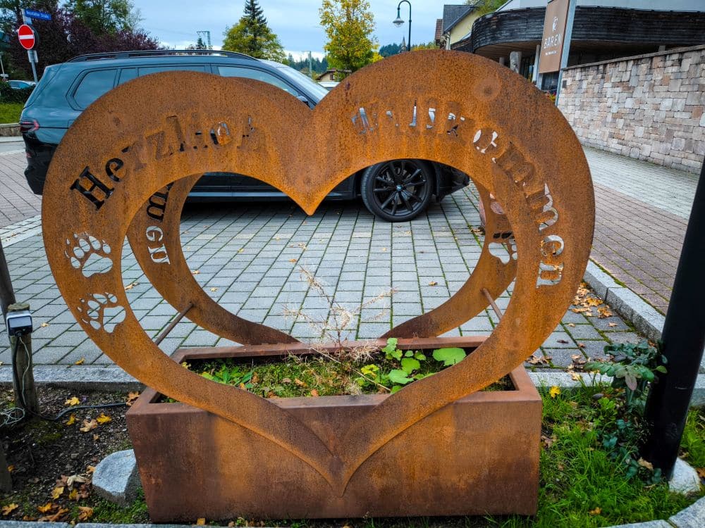Ein rostiges, herzförmiges Metallschild mit Pfotenabdrücken und der Aufschrift "Herzlich Willkommen" steht auf einem Blumenkasten in Titisee, vor einem geparkten Auto auf einer Kopfsteinpflasterstraße. Bäume und Gebäude umrahmen die charmante Szene.