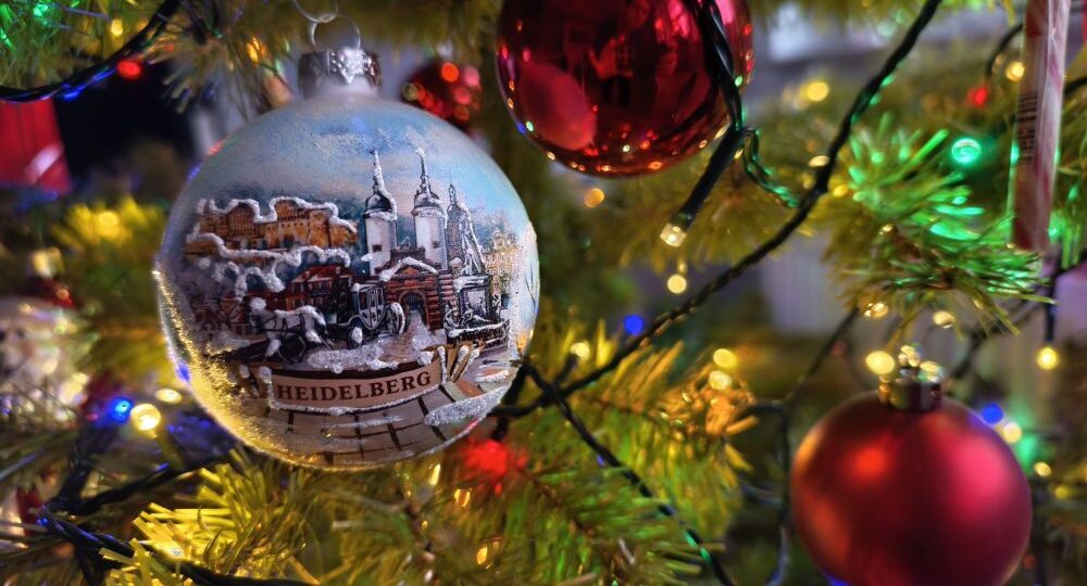 Ein geschmückter Weihnachtsbaum mit bunten Lichtern und Ornamenten, darunter eine gemalte Kugel mit einer verschneiten Heidelberger Szene, und rot glänzende Kugeln, die die nahe Umgebung widerspiegeln, bieten eine kleine Auszeit und wünschen fröhliche Weihnachten.