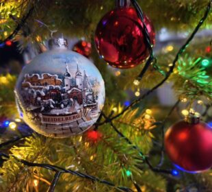 Ein geschmückter Weihnachtsbaum mit bunten Lichtern und Ornamenten, darunter eine gemalte Kugel mit einer verschneiten Heidelberger Szene, und rot glänzende Kugeln, die die nahe Umgebung widerspiegeln, bieten eine kleine Auszeit und wünschen fröhliche Weihnachten.