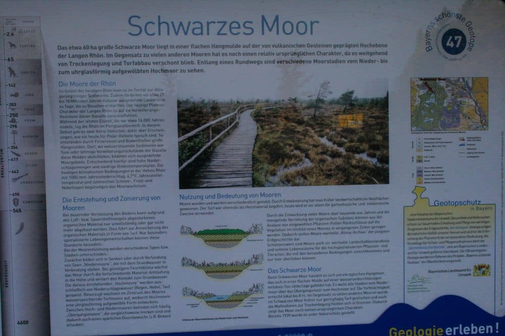 Eine Informationstafel über das Schwarze Moor enthält einen deutschen Text, Diagramme zur Moorbildung, eine Karte und ein Foto eines Holzstegs, der durch die grasbewachsene, feuchte Moorlandschaft unter grauem Himmel führt.