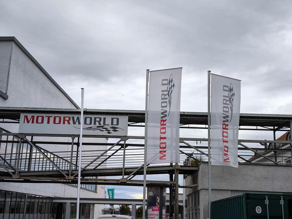 Drei MOTORWORLD-Schilder, darunter zwei senkrechte Fahnen und ein waagerechtes Schild, sind auf einer Fußgängerbrücke aus Metall und an Masten vor einem modernen Gebäude in Böblingen unter einem bewölkten Himmel angebracht.