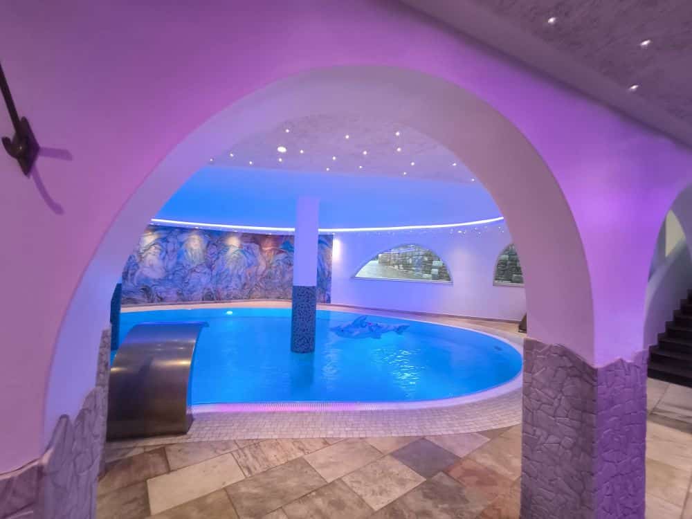 Ein luxuriöser Innenpool im Kastel verfügt über geschwungene Bögen, violette Beleuchtung, Mosaikfliesen, einen Wasserfall und ein dekoratives Wandgemälde an der Rückwand. Auf der rechten Seite ist eine Treppe zu sehen.