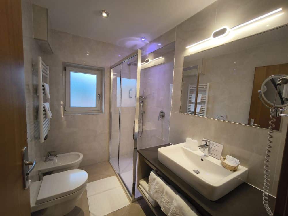 Modernes Kastel-Bad mit Glasdusche, wandmontiertem Waschbecken, Bidet und Toilette. Die Handtücher liegen gefaltet auf einem Regal unter dem Waschbecken, während die Toilettenartikel ordentlich auf der Arbeitsplatte liegen. Ein Milchglasfenster erfüllt den Raum mit natürlichem Licht.