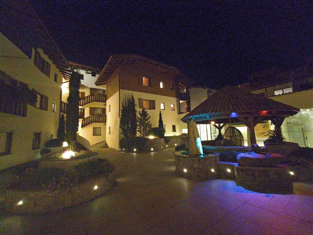 Eine nächtliche Hofszene in Kastel mit beleuchteten Wegen, einem runden Pavillon und weißen Gebäuden mit Holzbalkonen und Fenstern. Sanfte Lichter akzentuieren die Landschaftsgestaltung und den Pavillon und schaffen eine ruhige und einladende Atmosphäre.