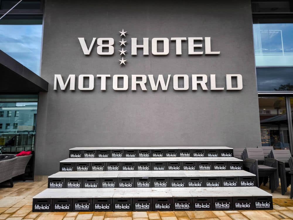 Großes V8 HOTEL MOTORWORLD-Schild an einer grauen Wand über gestapelten Kisten von fritz-kola in Böblingen, vor einem modernen Gebäude; rechts davon sind schwarze Stühle gestapelt.
