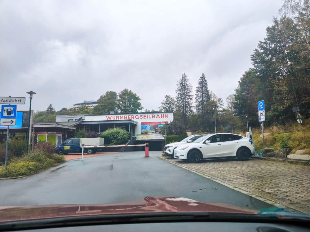 Blick aus dem Auto auf die Station der Wurmbergseilbahn in Braunlage an einem bewölkten Tag, mit geparkten weißen Autos, Bäumen, einem Parkschild und einer Straße, die zum Eingang führt.