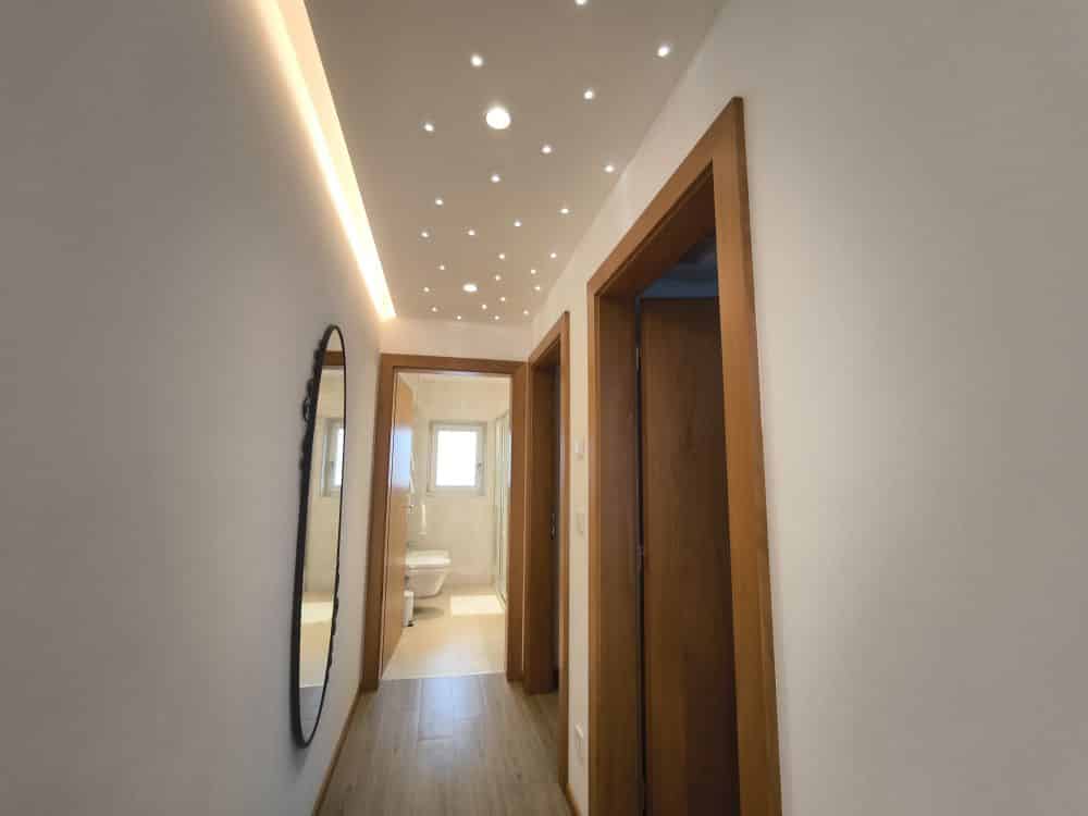 Ein Flur mit Kastel-Holzfußboden, weißen Wänden und zwei offenen Türen. An der Decke befinden sich kleine Einbaulampen und Umgebungslicht. Am Ende des Flurs ist ein Badezimmer mit einer Toilette und einem Fenster zu sehen. An der linken Wand hängt ein ovaler Spiegel.