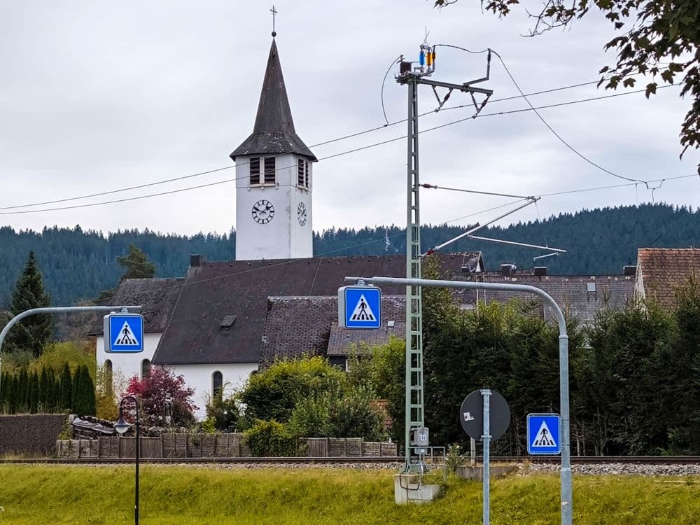 Eine weiße Kirche mit einem Glockenturm steht in Titisee hinter Bahnstromleitungen und mehreren Schildern für Fußgängerüberwege, umgeben von Bäumen und Häusern, mit einem bewaldeten Hügel im Hintergrund.