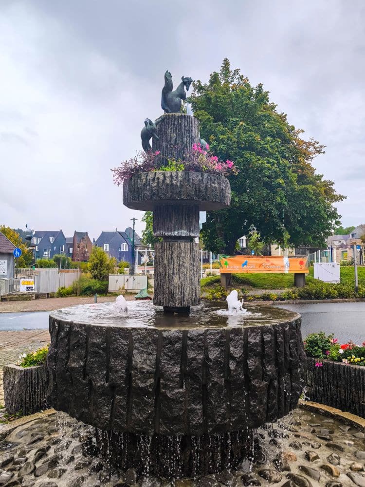 Auf einem gepflasterten Platz in Braunlage steht ein steinerner Brunnen, dessen Wasser von mehreren Stufen fließt und der von Tierskulpturen und rosafarbenen Blumen gekrönt wird. Im Hintergrund sind Bäume, Gebäude und ein bewölkter Himmel zu sehen.