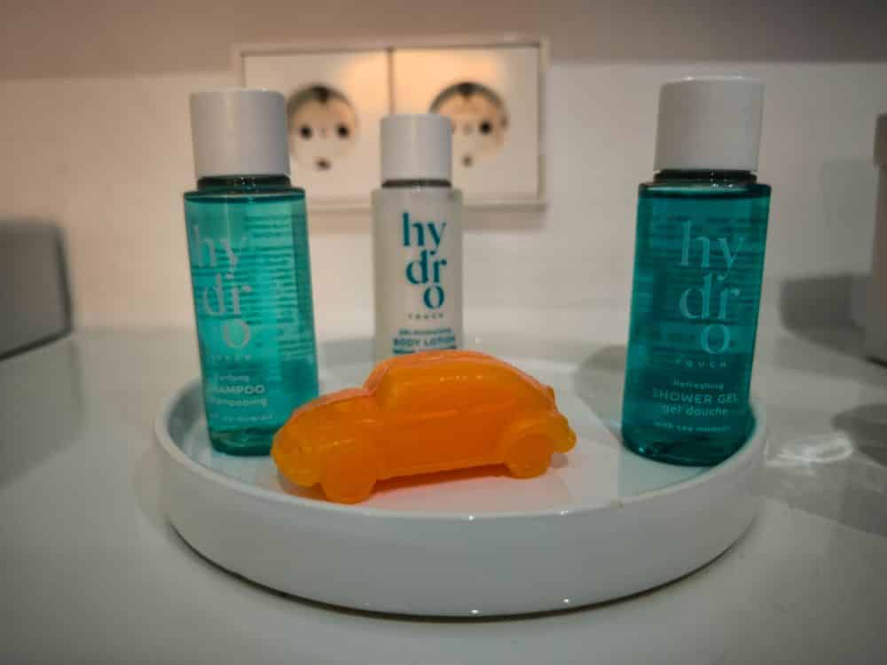 Drei kleine Toilettenflaschen mit der Aufschrift "Hydro" und eine orangefarbene Seife in Form eines Autos stehen auf einem weißen Tablett in einem Böblinger Hotelzimmer, im Hintergrund ist eine Steckdose zu sehen. Die Flaschen enthalten Shampoo, Körperlotion und Duschgel.