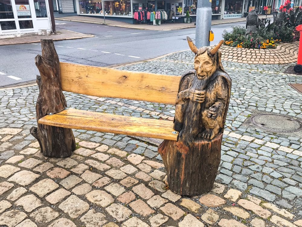 Eine hölzerne Sitzbank im Freien in Braunlage, an deren einem Ende eine detaillierte Figur eines gehörnten Wesens mit einem Becher in der Hand geschnitzt ist, steht auf einem Kopfsteinpflaster in der Nähe einer Straßenecke mit Geschäften im Hintergrund.