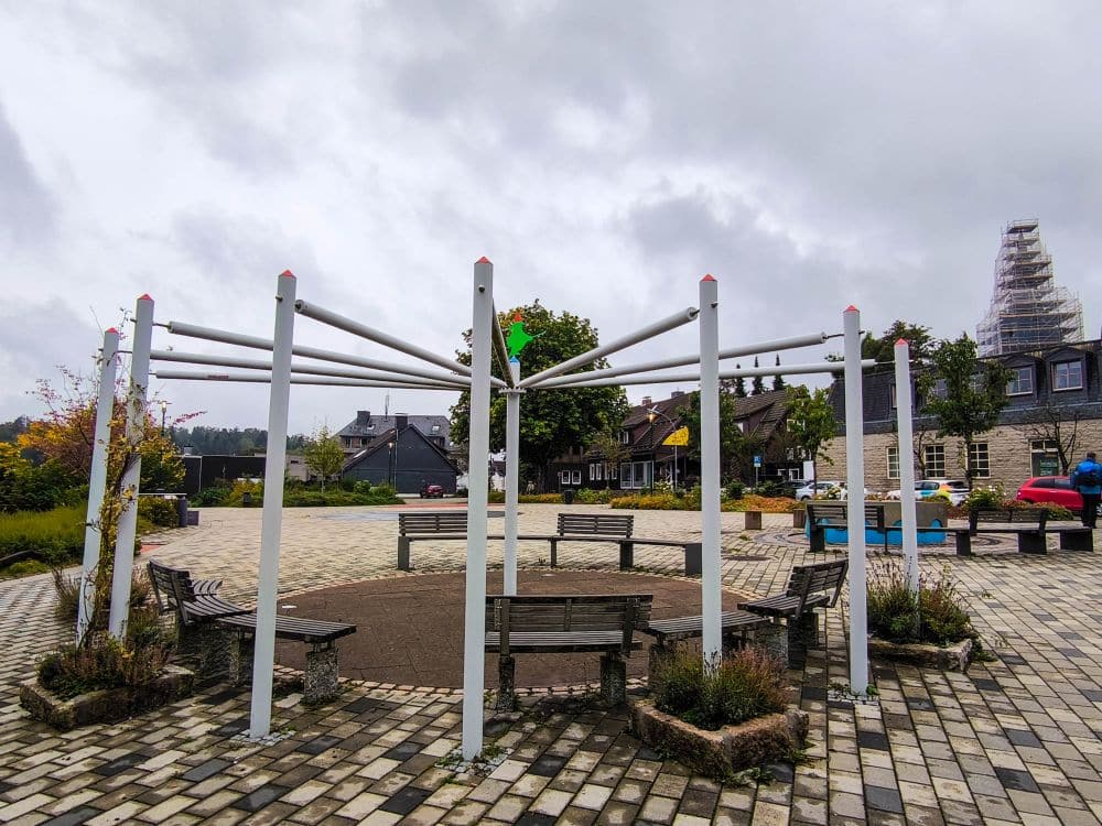 Ein kreisrunder Sitzplatz mit Bänken und Metallstangen steht in der Mitte eines gepflasterten Platzes in Braunlage, umgeben von Pflanzen und Gebäuden unter einem bewölkten Himmel.