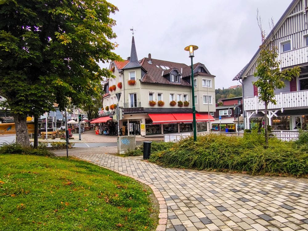 Ein gepflasterter Gehweg schlängelt sich über den kleinen Stadtplatz von Braunlage mit grünem Gras, Bäumen und einer Straßenlaterne. An der Kreuzung steht an einem bewölkten Tag ein dreistöckiges Gebäude mit roten Markisen und Blumenkästen.
