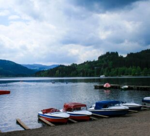 Ruderboote mit farbenfrohen Verkleidungen liegen am Ufer des Titisees, eines ruhigen Sees, der von Hügeln und dichten grünen Wäldern umgeben ist, unter einem bewölkten Himmel. Ein paar Leute stehen auf einem nahe gelegenen Holzsteg.