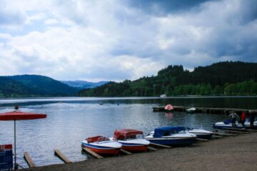 Ruderboote mit farbenfrohen Verkleidungen liegen am Ufer des Titisees, eines ruhigen Sees, der von Hügeln und dichten grünen Wäldern umgeben ist, unter einem bewölkten Himmel. Ein paar Leute stehen auf einem nahe gelegenen Holzsteg.