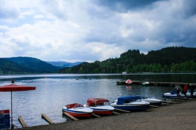 Ruderboote mit farbenfrohen Verkleidungen liegen am Ufer des Titisees, eines ruhigen Sees, der von Hügeln und dichten grünen Wäldern umgeben ist, unter einem bewölkten Himmel. Ein paar Leute stehen auf einem nahe gelegenen Holzsteg.