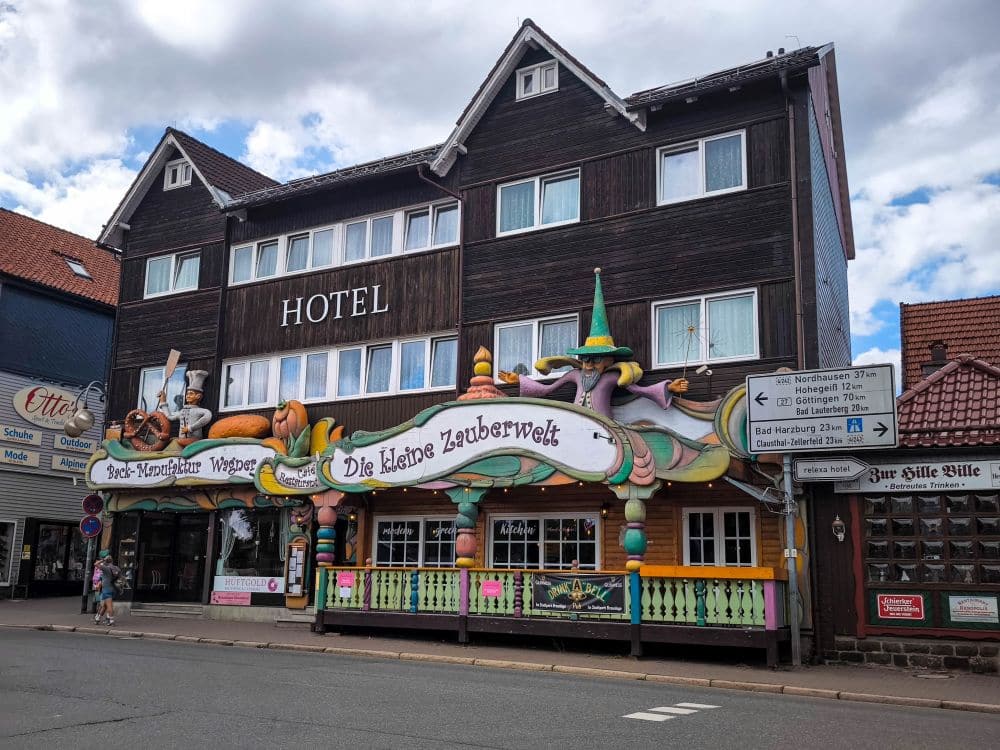 Ein skurriler, farbenfroher Laden mit fantasievollen Dekorationen und einem Schild mit der Aufschrift "Die kleine Zauberwelt" bringt Charme in eine ruhige Straße in Braunlage, die sich an ein großes, dunkles Holzhotel mit weiß gerahmten Fenstern schmiegt.