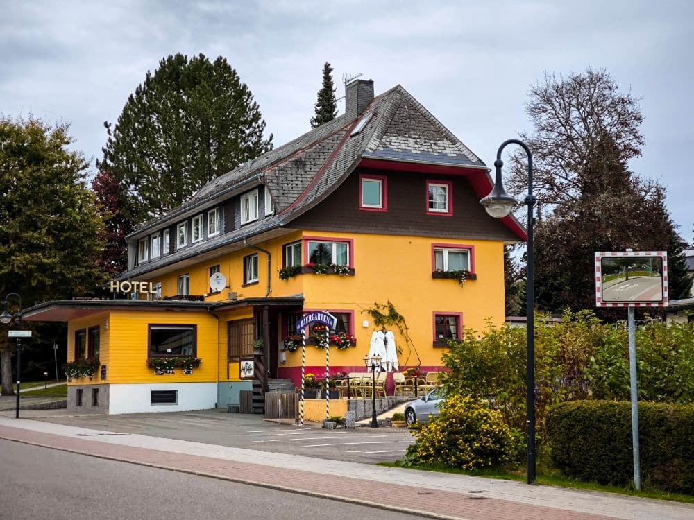 Ein gelb-braunes Hotel mit Blumenkästen steht an einer Straßenecke in Titisee, umgeben von Grün und kleinen Bäumen unter einem bewölkten Himmel. Im Vordergrund sind ein Straßenschild und ein Laternenpfahl zu sehen.