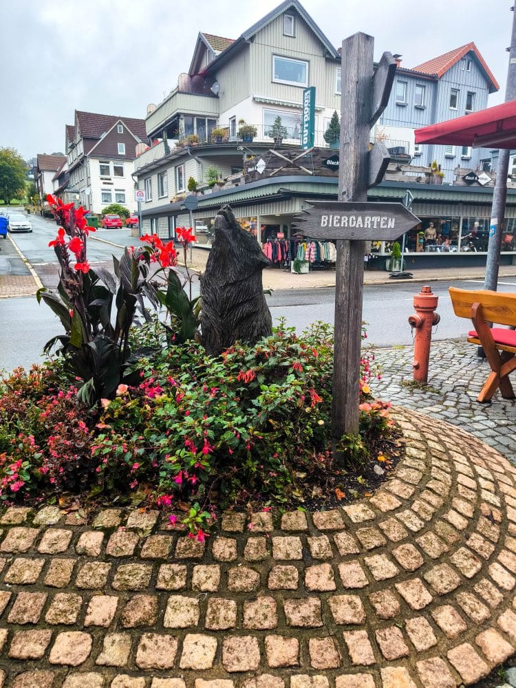 Auf einem Kreisverkehr mit Kopfsteinpflaster in Braunlage stehen Blumen, eine hölzerne Bärenskulptur und ein "Biergarten"-Schild. Im Hintergrund sind unter dem wolkenverhangenen Himmel Geschäfte und Häuser zu sehen.