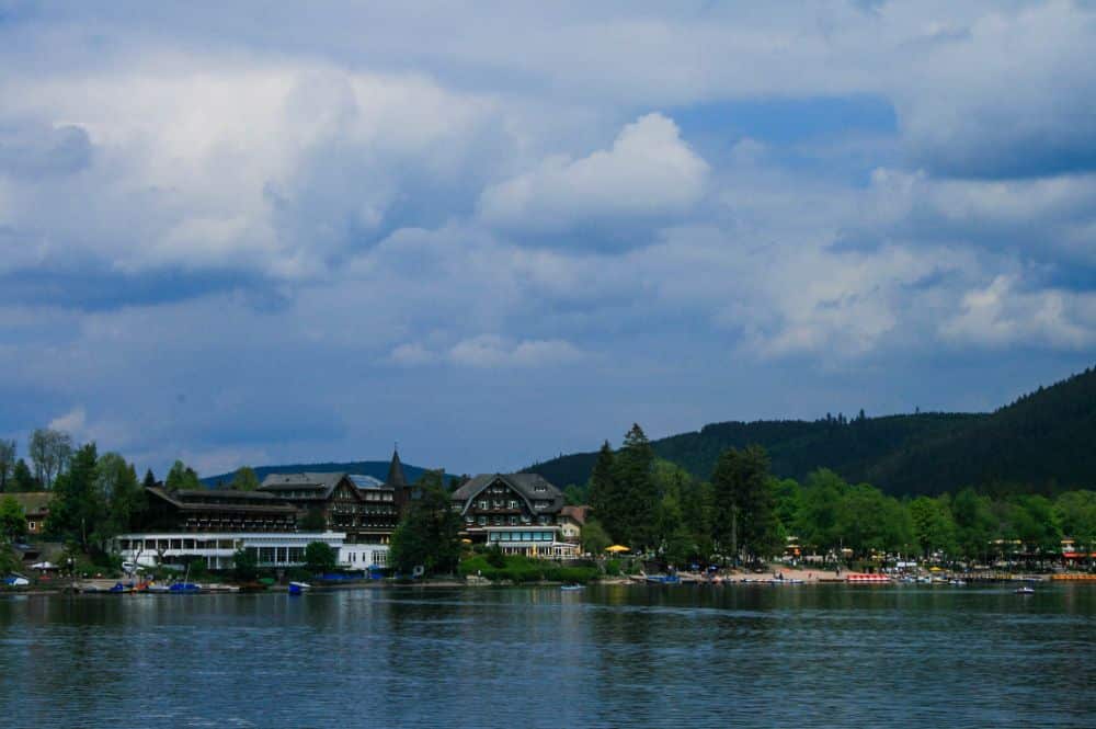 Eine Seeansicht mit Gebäuden und Bäumen unter einem teilweise bewölkten Himmel, mit Hügeln und Wäldern im Hintergrund - das ruhige Wasser des Titisees spiegelt die atemberaubende Landschaft im Vordergrund wunderbar wider.