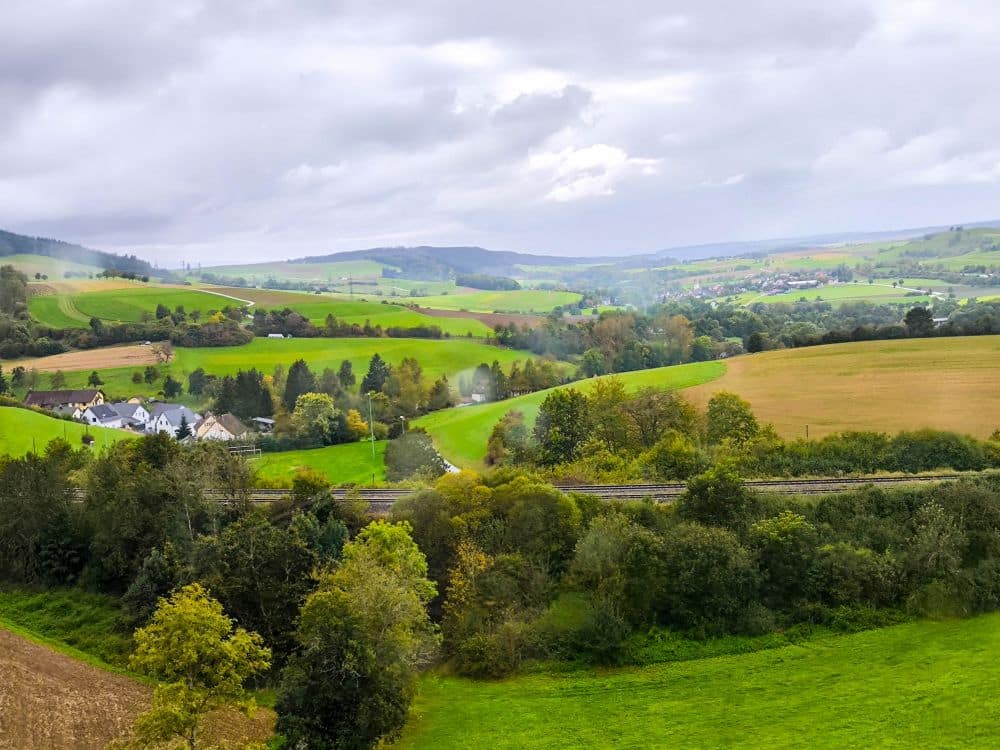 Eine malerische Landschaft mit sanften grünen Hügeln, vereinzelten Bäumen, ein paar Häusern und einer Eisenbahnlinie, die sich unter einem wolkenverhangenen Himmel durch die Titisee-Landschaft schlängelt.