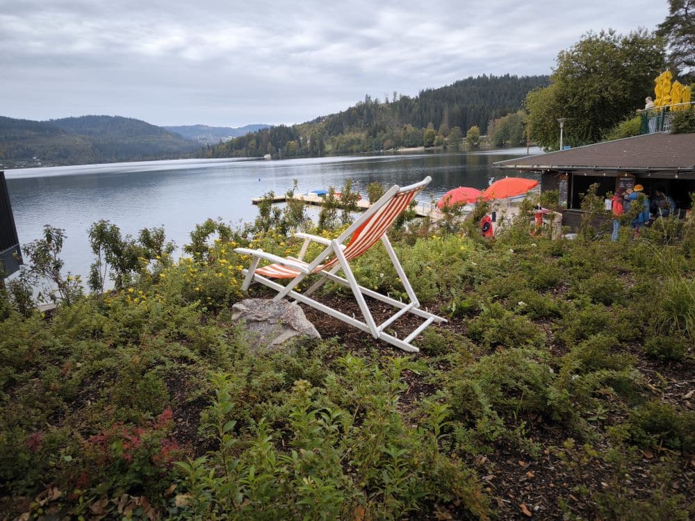 Ein großer weiß-orange gestreifter Liegestuhl steht auf einem grasbewachsenen Hügel mit Blick auf den Titisee, mit bewaldeten Hügeln im Hintergrund. In der Nähe eines Gebäudes am Ufer des Sees sind Menschen und rote Regenschirme zu sehen.