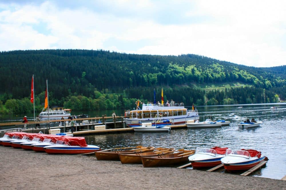 Ruder- und Tretboote liegen aufgereiht am Sandstrand des Titisees, mit einem Steg und mehreren größeren Booten auf dem ruhigen See. Im Hintergrund erhebt sich ein bewaldeter Hügel unter einem teilweise bewölkten Himmel.