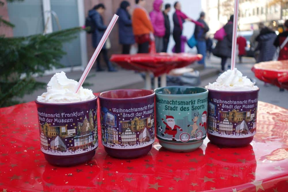 Vier festliche Weihnachtsbecher, zwei davon mit Schlagsahne und Strohhalm, stehen auf einem Tisch mit rotem Sternmuster. Im verschwommenen Hintergrund stehen Menschen in Winterkleidung auf einem Markt im Freien und unterhalten sich.