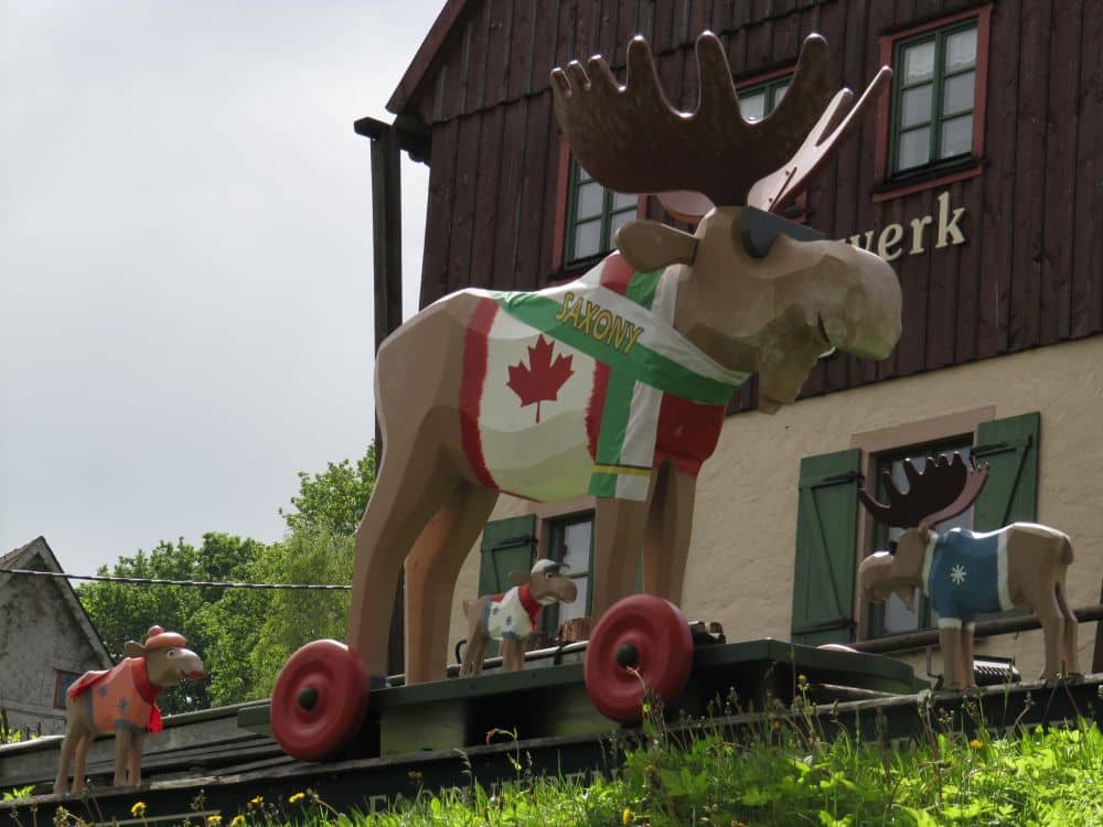 Drei große hölzerne Elchstatuen mit Rädern stehen auf einem Dach und sind mit verschiedenen Fahnen und Schals geschmückt. Eine von ihnen trägt eine kanadische Flagge an der Seite, was eine festliche Weihnachtsstimmung hervorruft. Ein Gebäude mit grünen Fenstern bildet die Kulisse.
