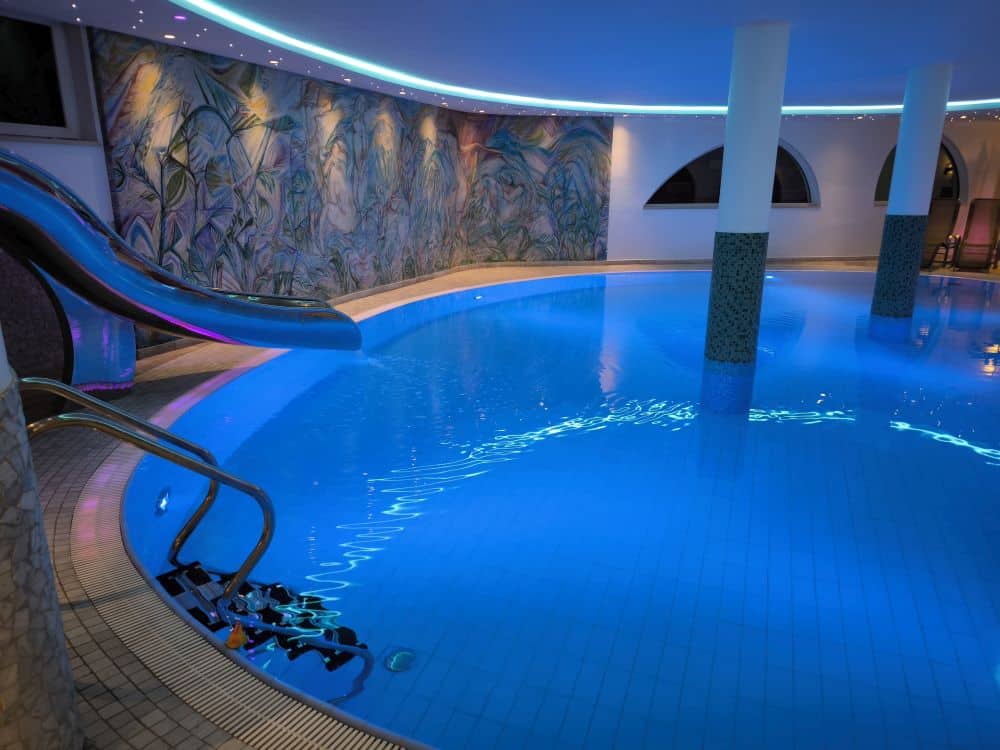 Hallenbad mit blauer Beleuchtung, einer geschwungenen Wasserrutsche und gefliesten Wänden. Die Unterwassertreppe, die dekorative Wandmalerei an einer Wand und die gewölbten Fenster sind im Kastel-Design gehalten. Die Umgebung wirkt ruhig und einladend.